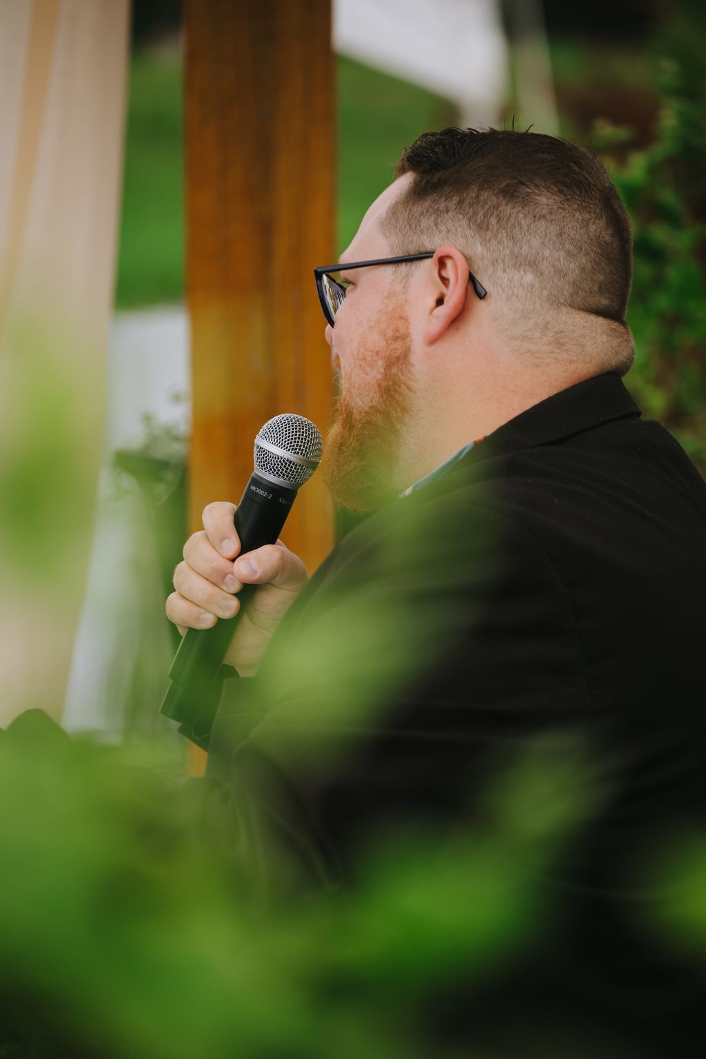 Groom speaking into microphone outdoors with blurred green background during wedding ceremony