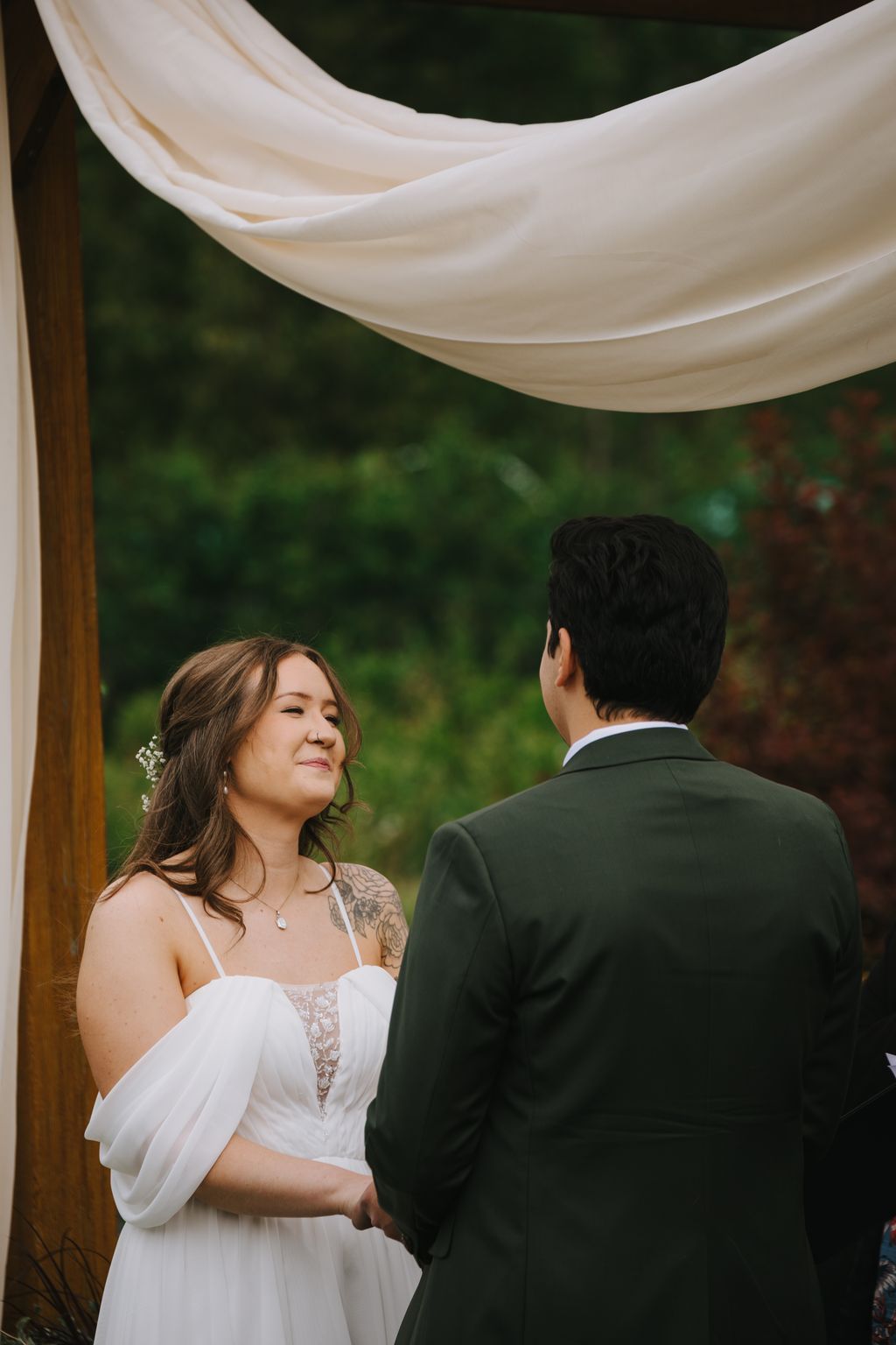 Bride and groom face each other under flowing white fabric canopy with lush green backdrop during outdoor ceremony