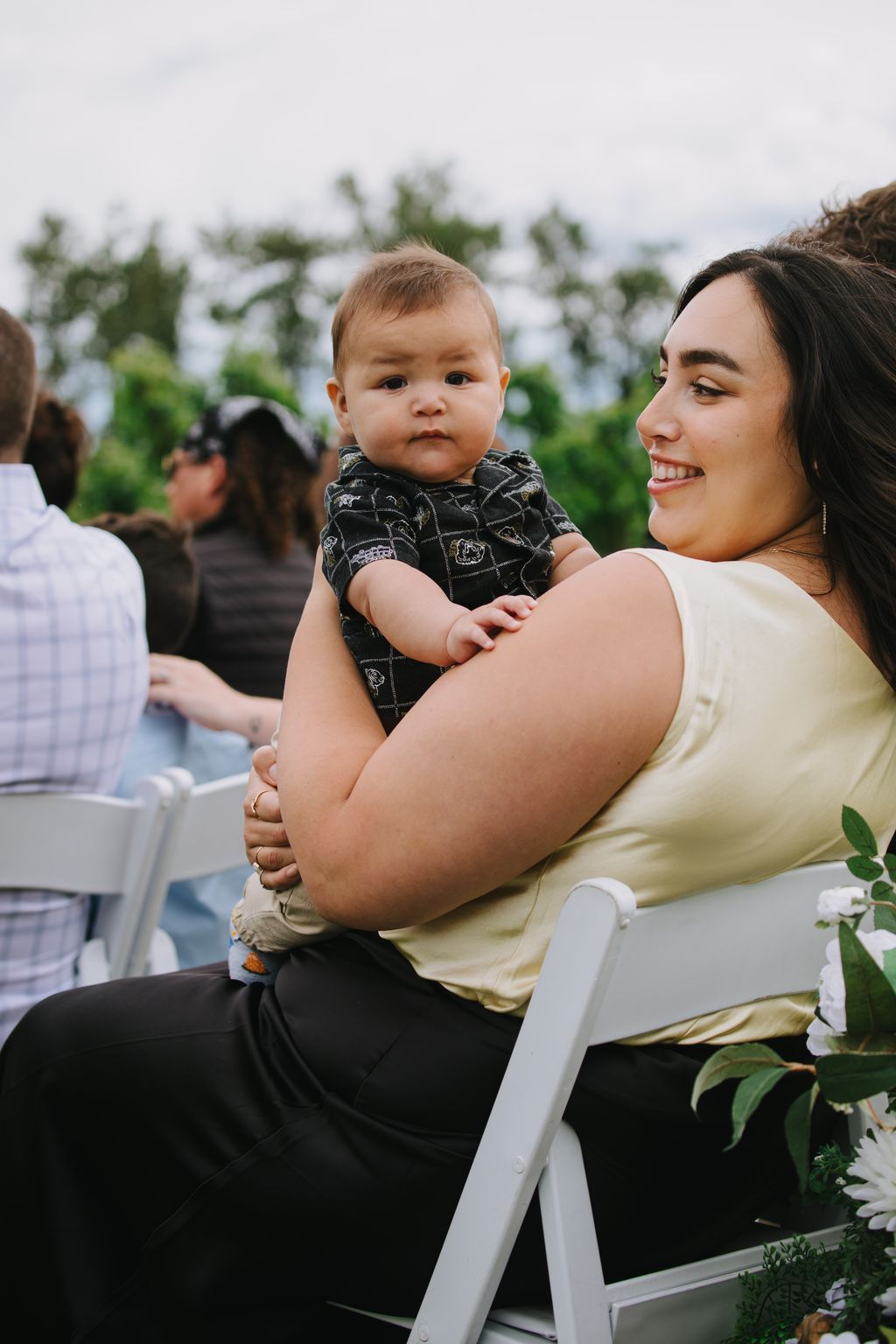 Infant in black patterned shirt held by smiling parent outdoors, soft natural light with blurred green foliage background