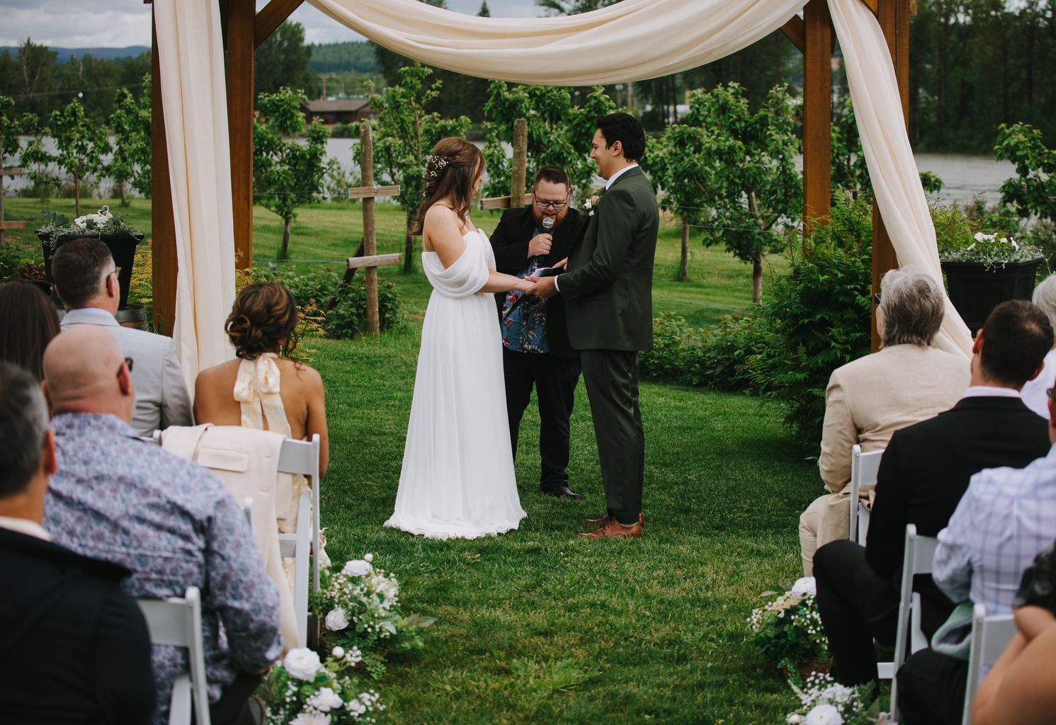 Bride in white flowing gown and groom in olive suit exchange rings during outdoor garden ceremony under draped fabric arbor