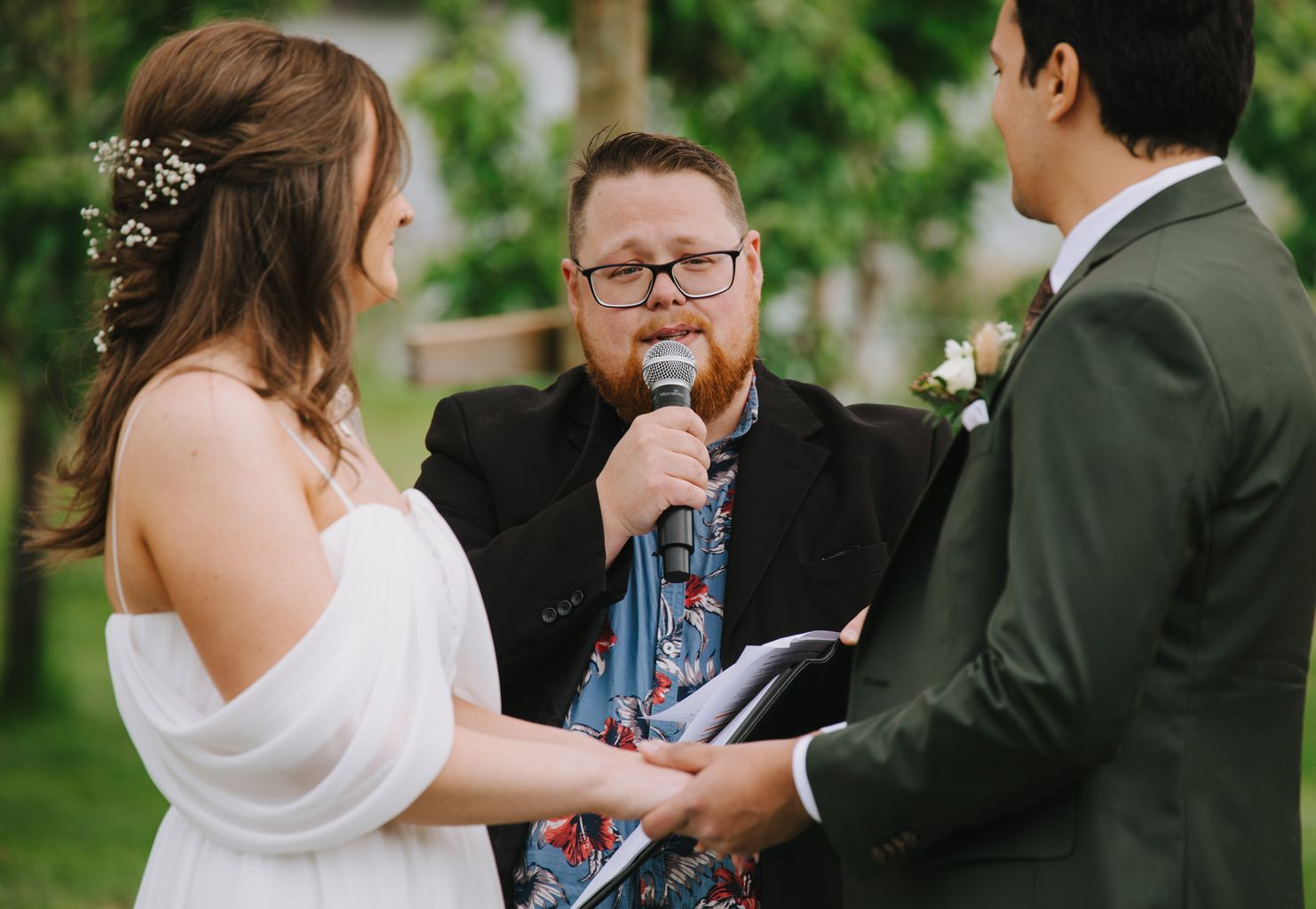 Man with beard and glasses speaks into microphone at outdoor event, holding papers, with lush greenery in background