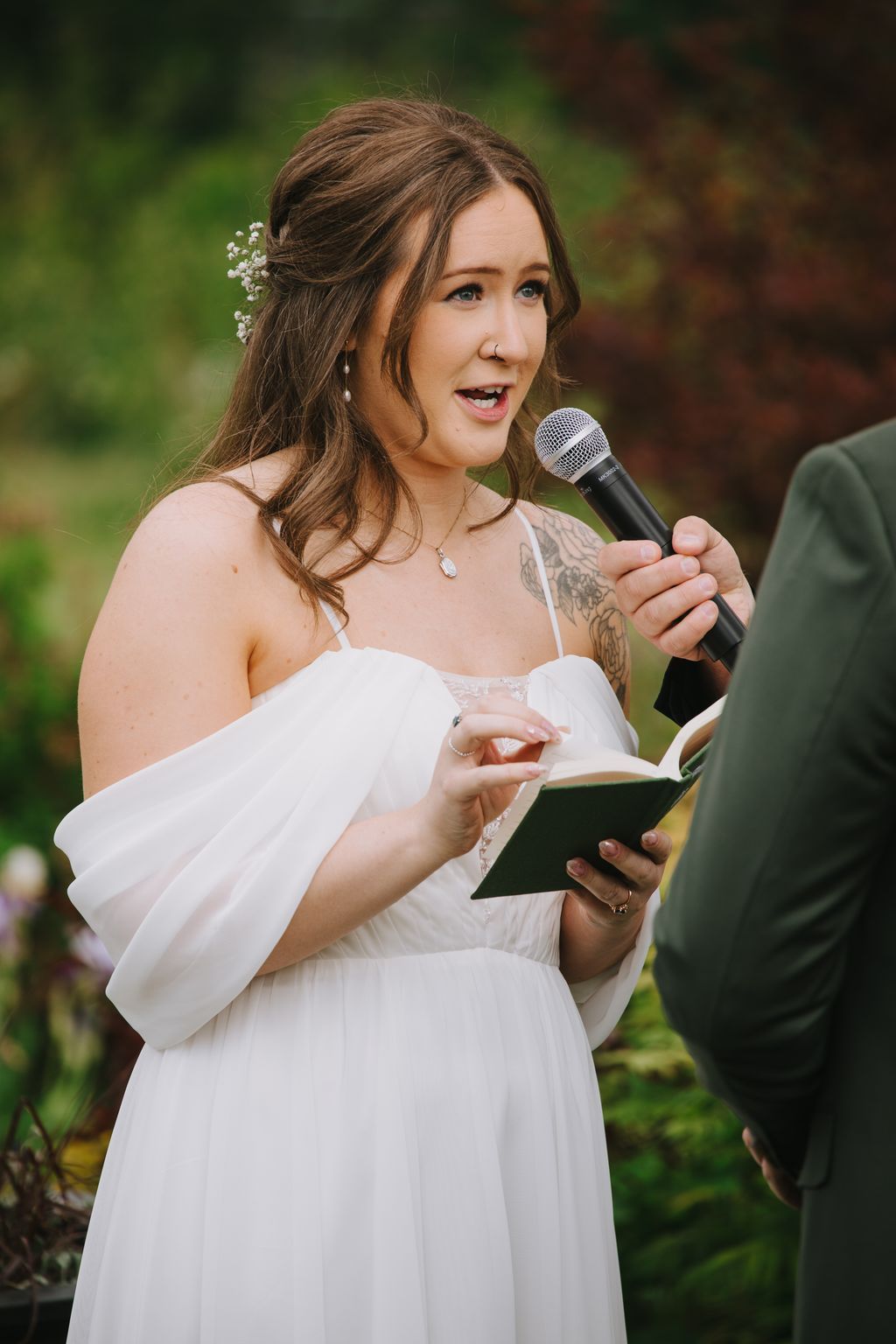 Bride in white dress holding microphone and reading from tablet during outdoor ceremony with baby''s breath in hair