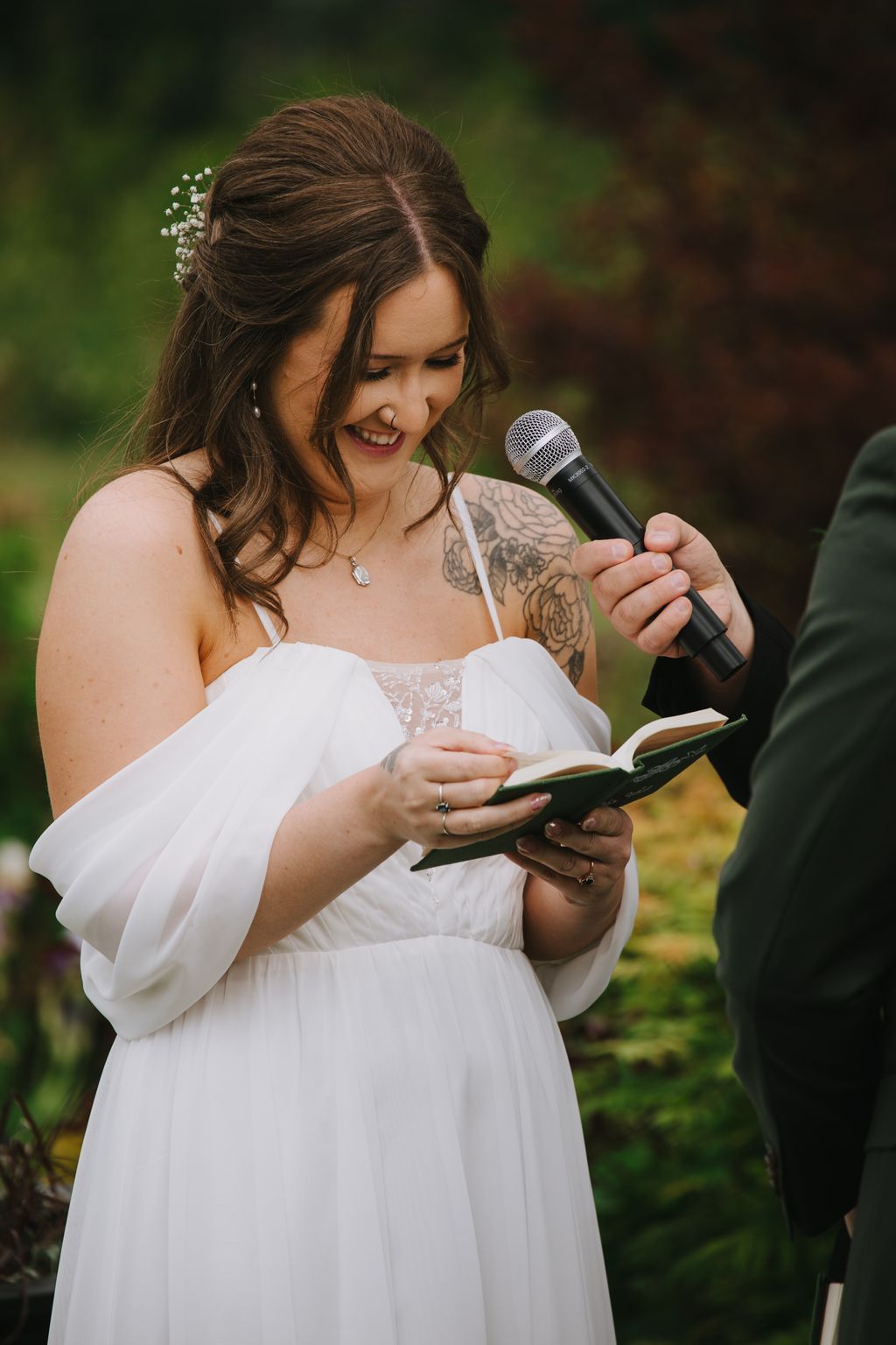 Bride in white dress reading vows from book while holding microphone, smiling outdoors with soft green background
