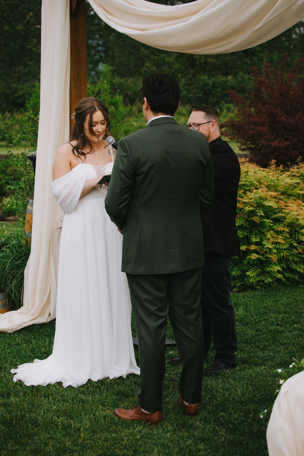 Bride in white dress exchanges vows with groom under cream fabric canopy during outdoor garden ceremony