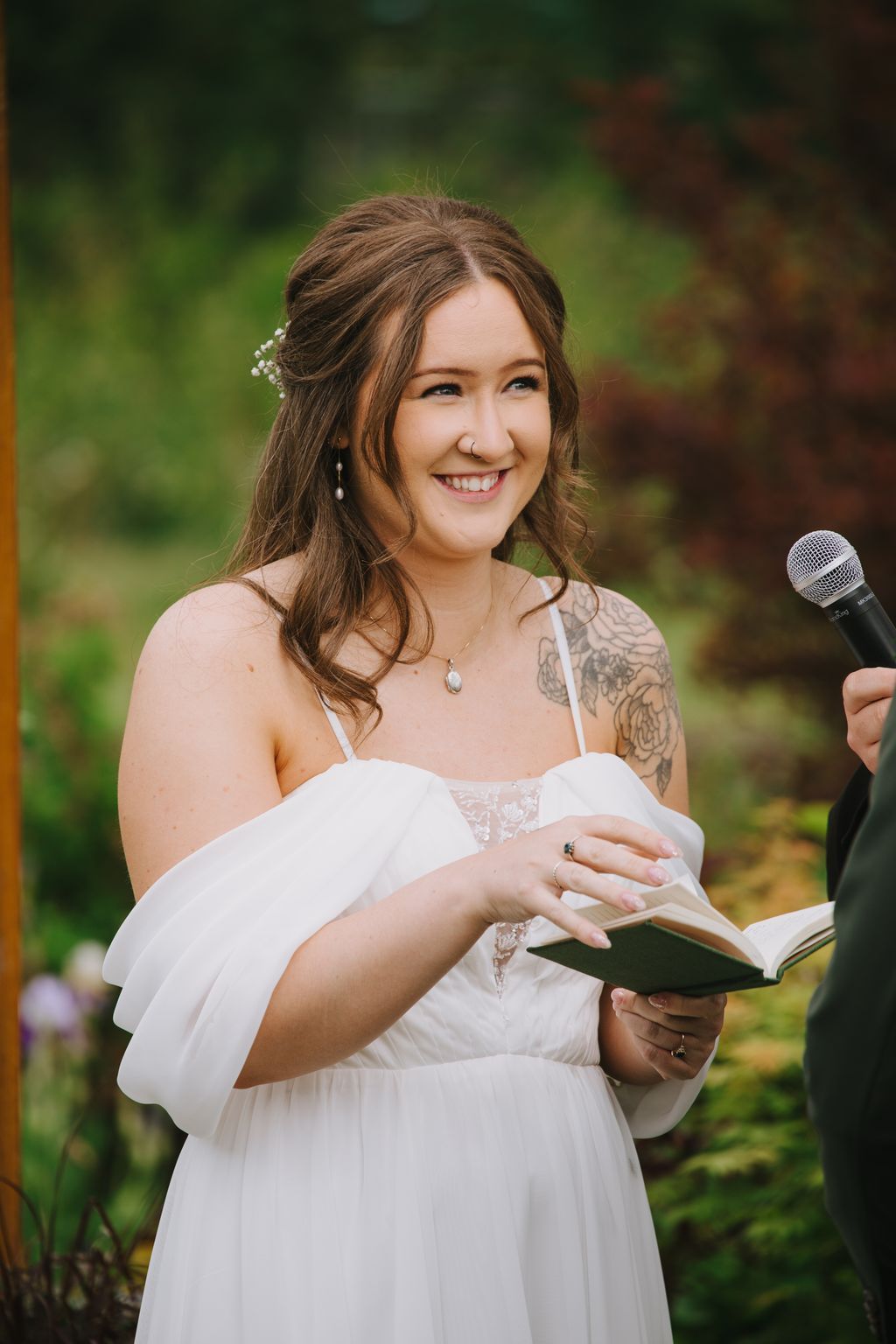 Smiling bride in white dress holding vows outdoors with soft green background and natural light