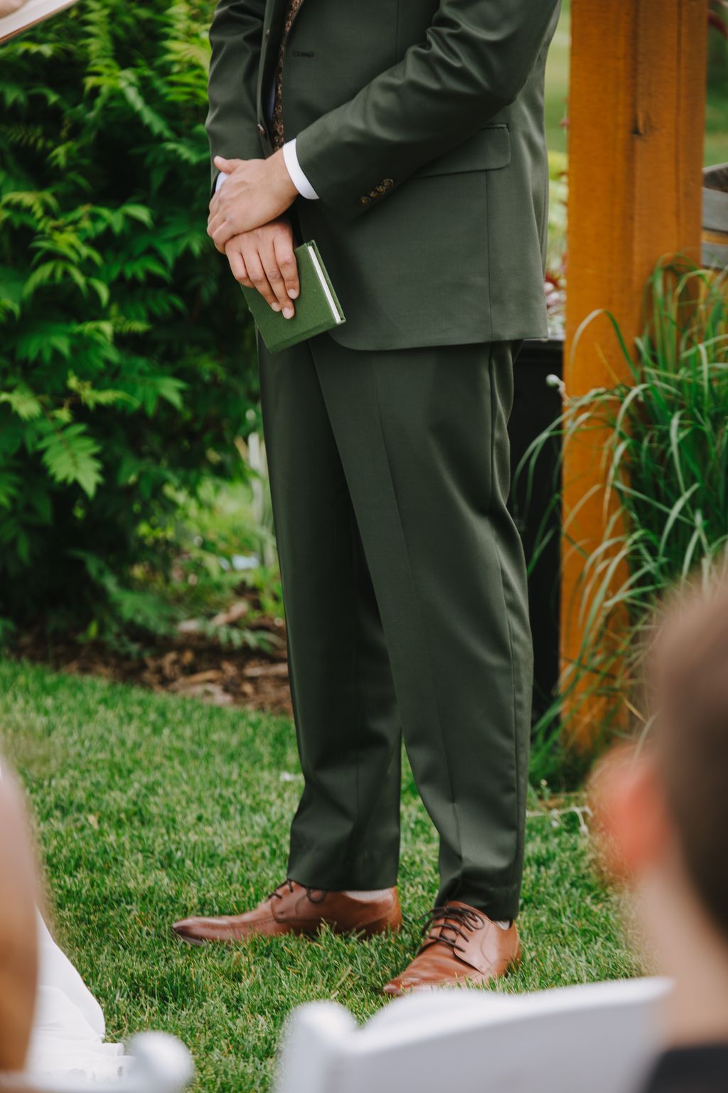 Groom in olive suit holding green clutch stands on grass during outdoor wedding ceremony
