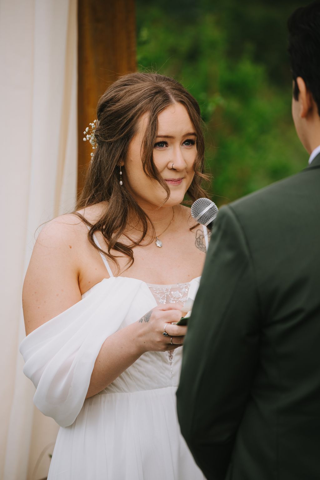 Bride in white dress holding microphone, speaking outdoors with soft green background and delicate baby''s breath in her hair