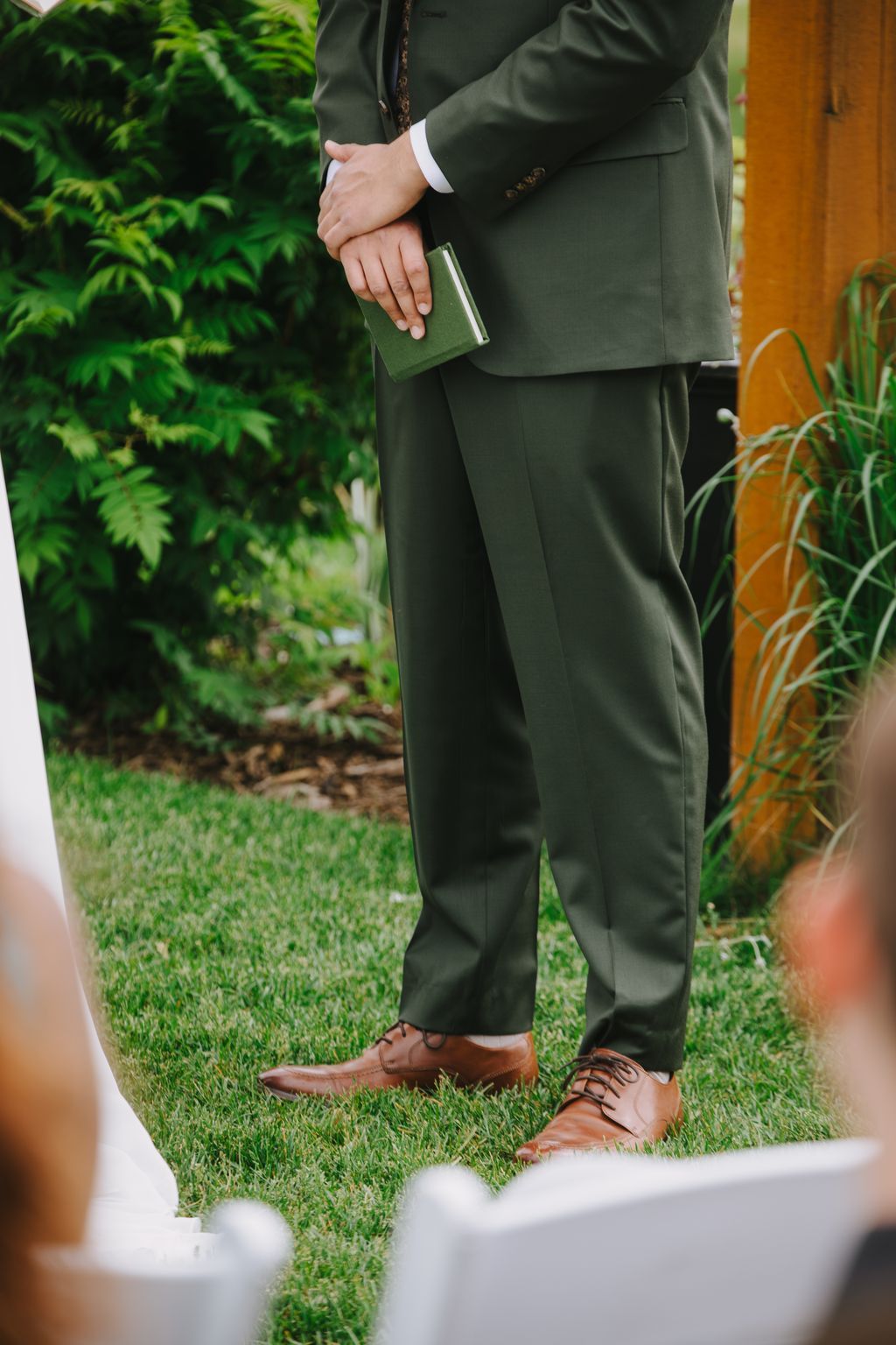 Groom in olive suit holding green clutch stands on grass with brown leather shoes visible, outdoor garden wedding detail shot