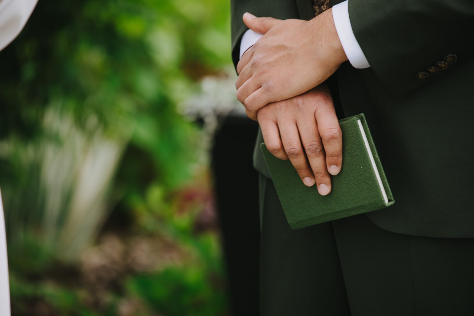 Couple''s hands clasped together outdoors with soft green foliage blurred in background
