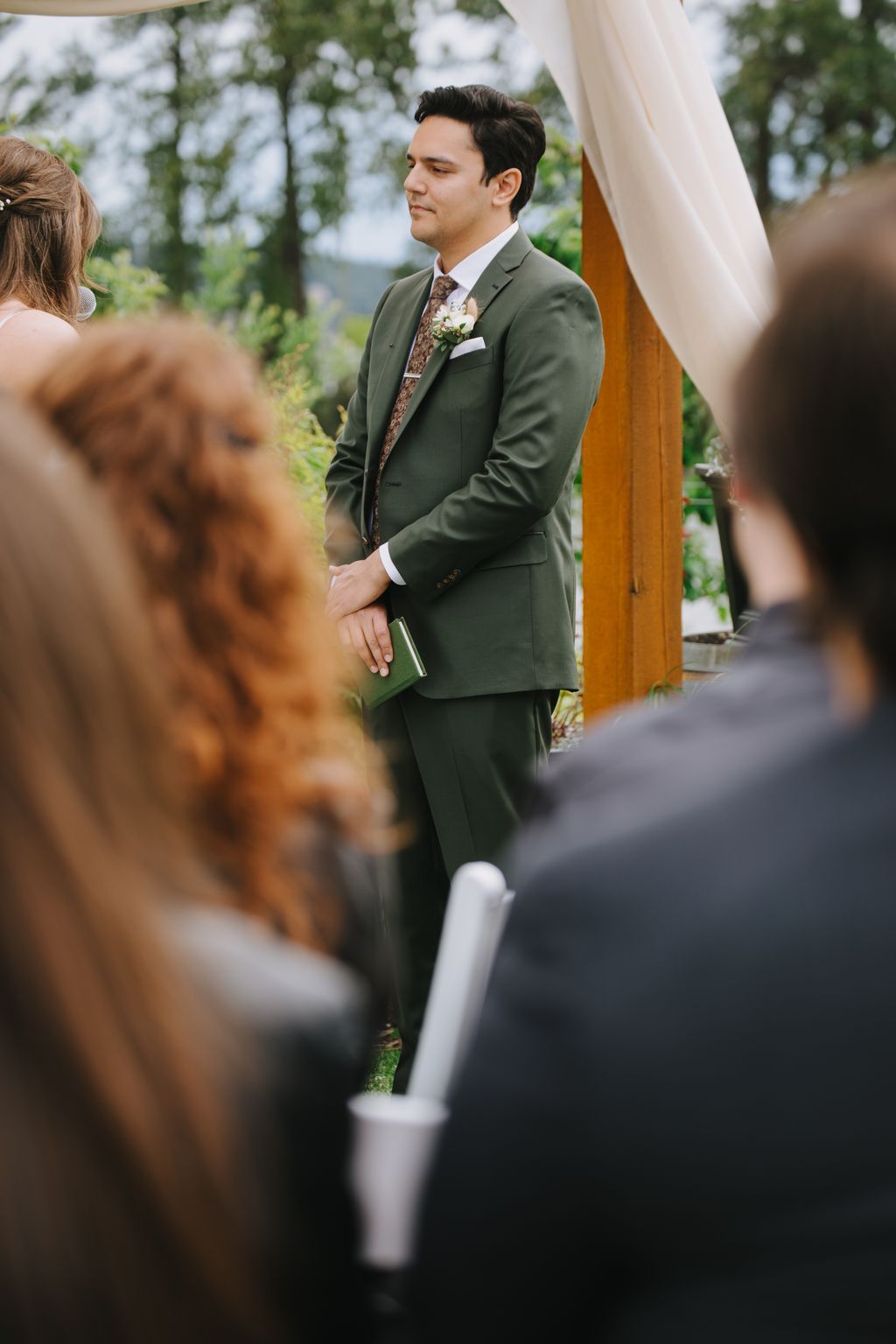 Groom in olive suit with floral tie and boutonniere stands at altar during outdoor wedding ceremony, holding green program, with soft natural light and trees…