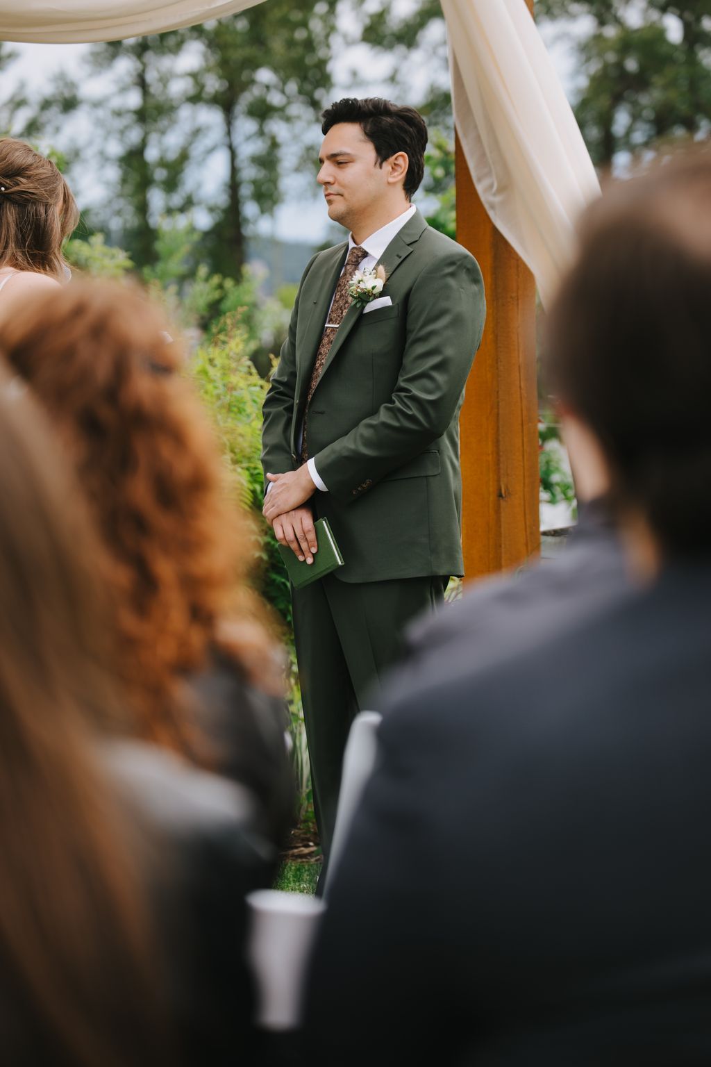 Groom in sage green suit with floral tie and boutonniere stands under sheer fabric canopy during outdoor wedding ceremony