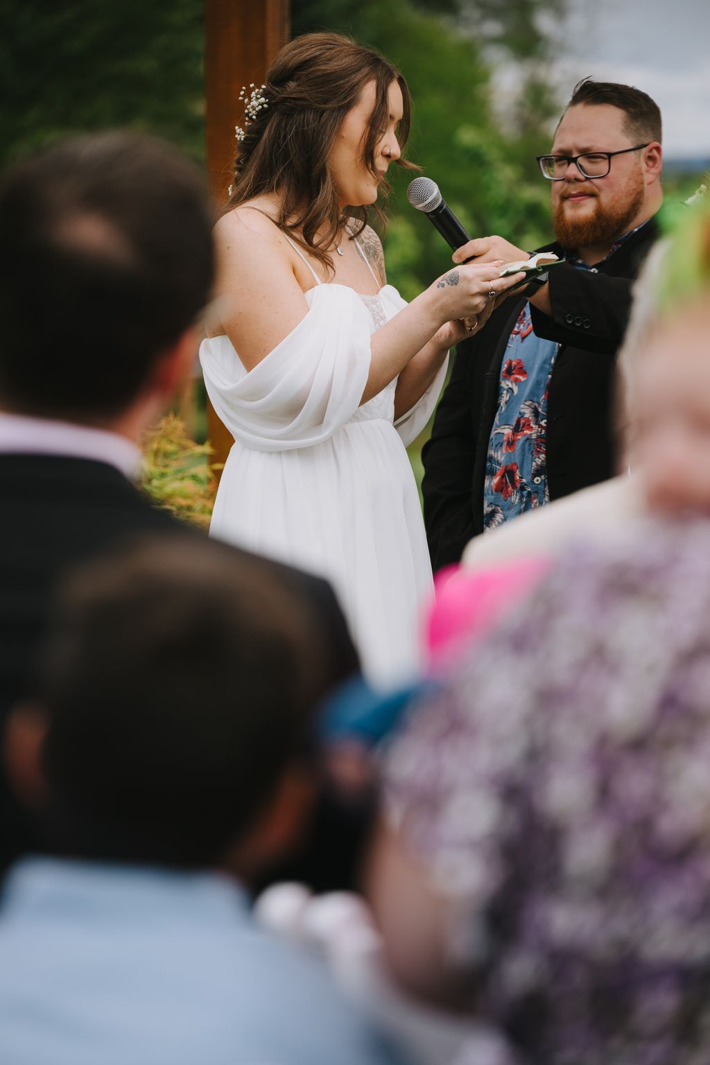 Bride in white flowing dress speaks at microphone with baby''s breath in her hair during outdoor garden ceremony