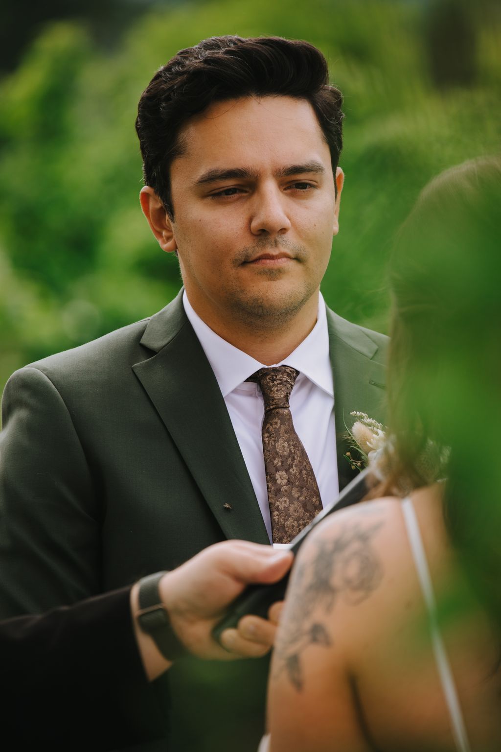 Groom in sage green suit and patterned tie gazes downward during outdoor ceremony with blurred green background