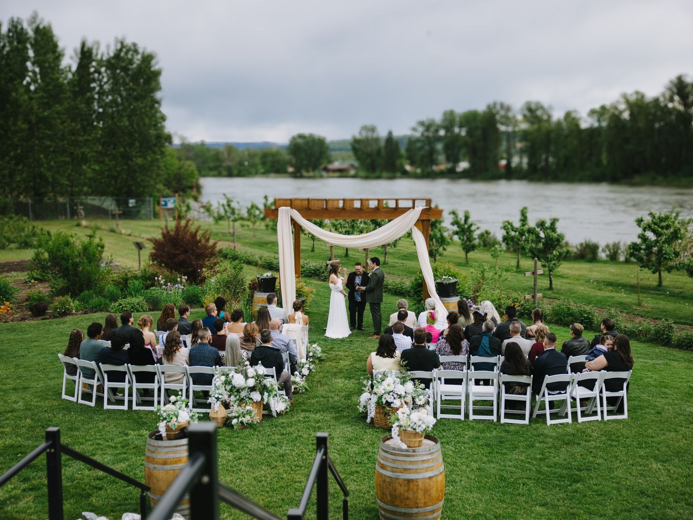 Bride and groom exchange vows under wooden arbor with white fabric at riverside garden ceremony surrounded by guests and white floral arrangements