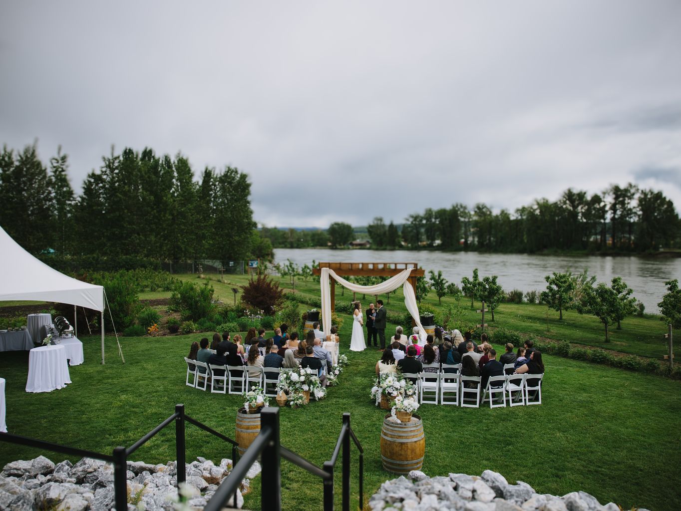 Bride and groom exchange vows beneath wooden arbor draped in white fabric, guests seated on lawn overlooking river under cloudy sky