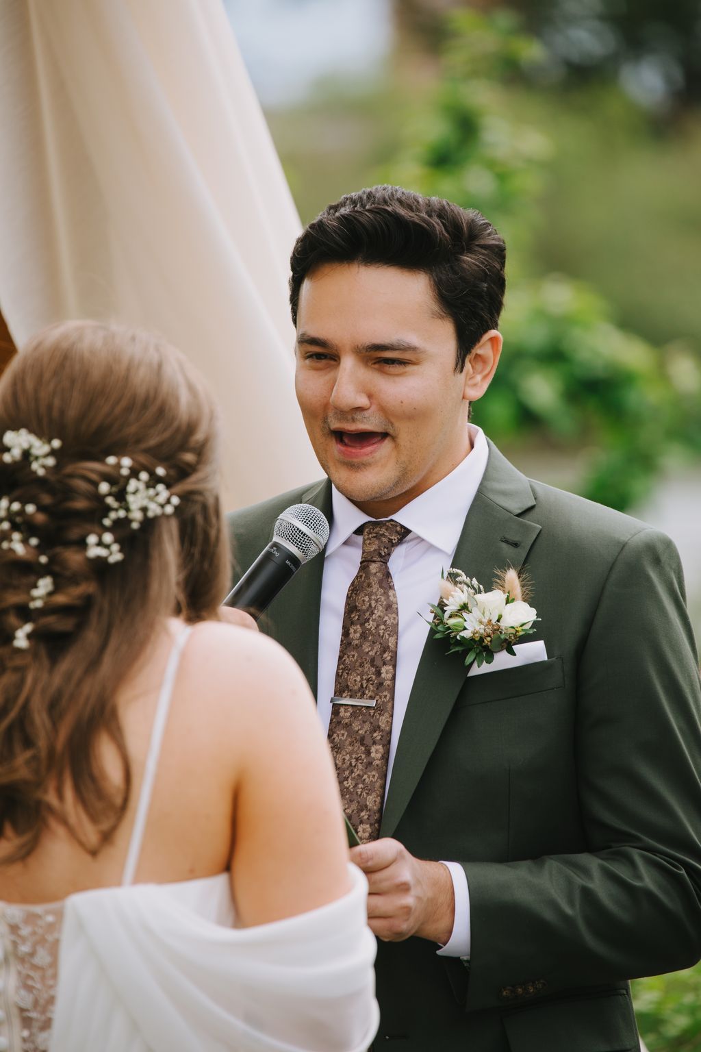 Groom in sage green suit and patterned tie speaks into microphone during outdoor wedding ceremony, smiling at bride