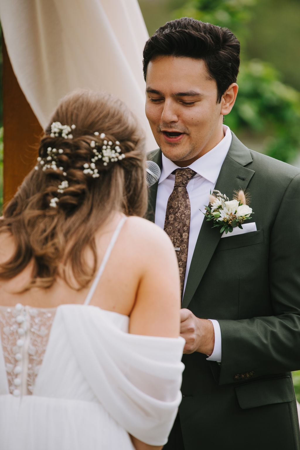 Groom speaks vows to bride during outdoor ceremony, bride''s hair adorned with baby''s breath flowers