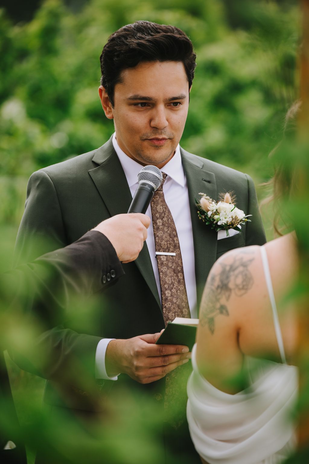 Groom in sage green suit and brown patterned tie holding microphone and vows during outdoor wedding ceremony with blurred green background