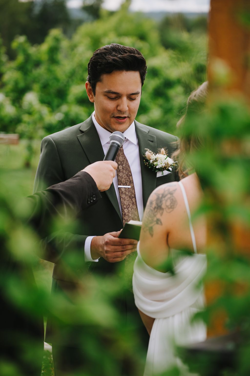 Groom in sage green suit reads vows to bride during outdoor garden ceremony with lush green backdrop