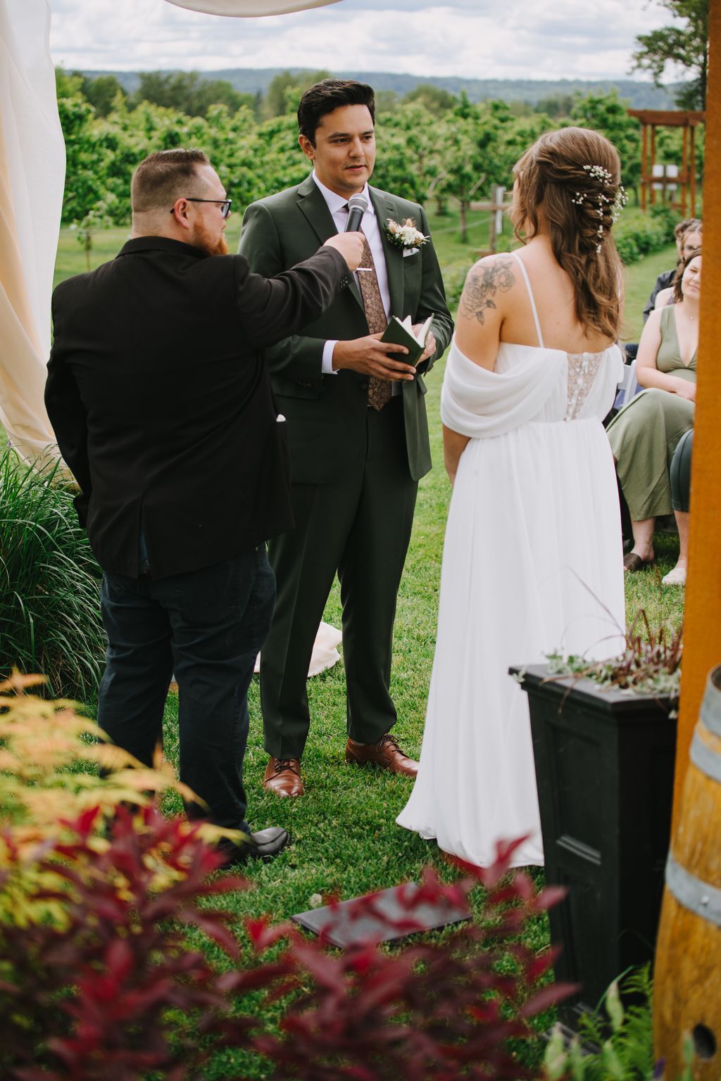 Groom in olive suit holds microphone during vineyard wedding ceremony with bride in white dress and officiant beside him