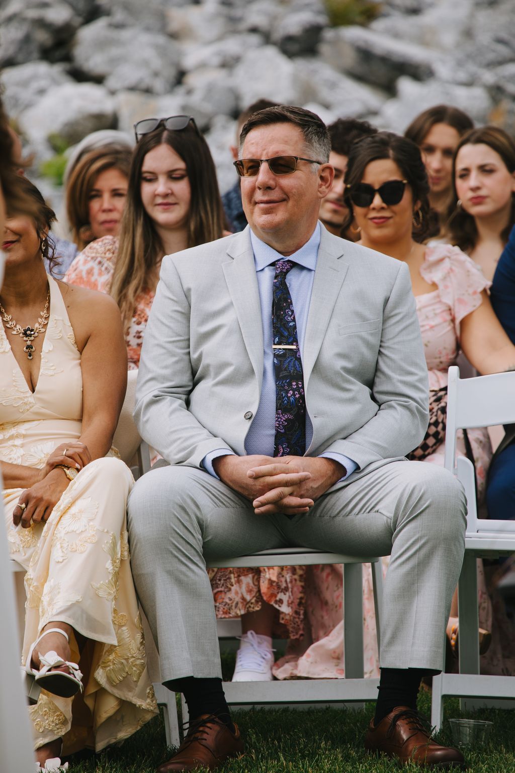 Groom in light gray suit and patterned tie sits smiling during outdoor ceremony with rocky backdrop and guests nearby