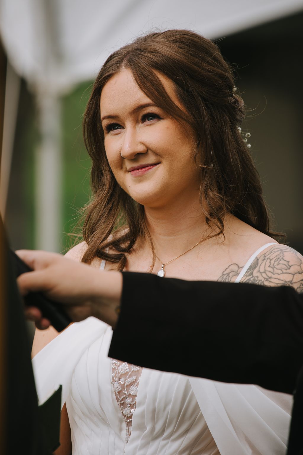 Bride in white lace dress with pendant necklace smiles softly outdoors against blurred green background