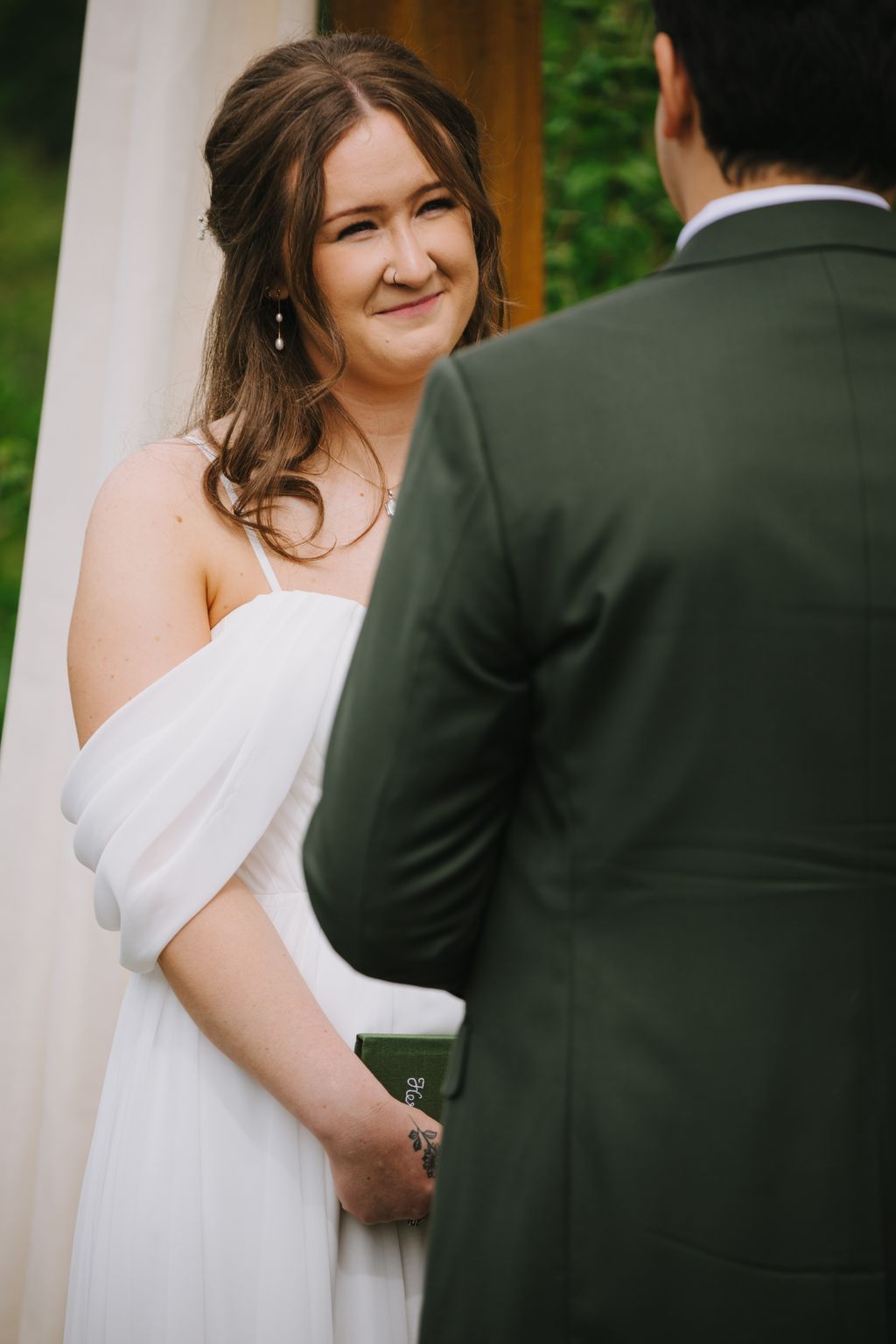 Bride in white dress smiles at groom in dark suit holding green ceremony book outdoors