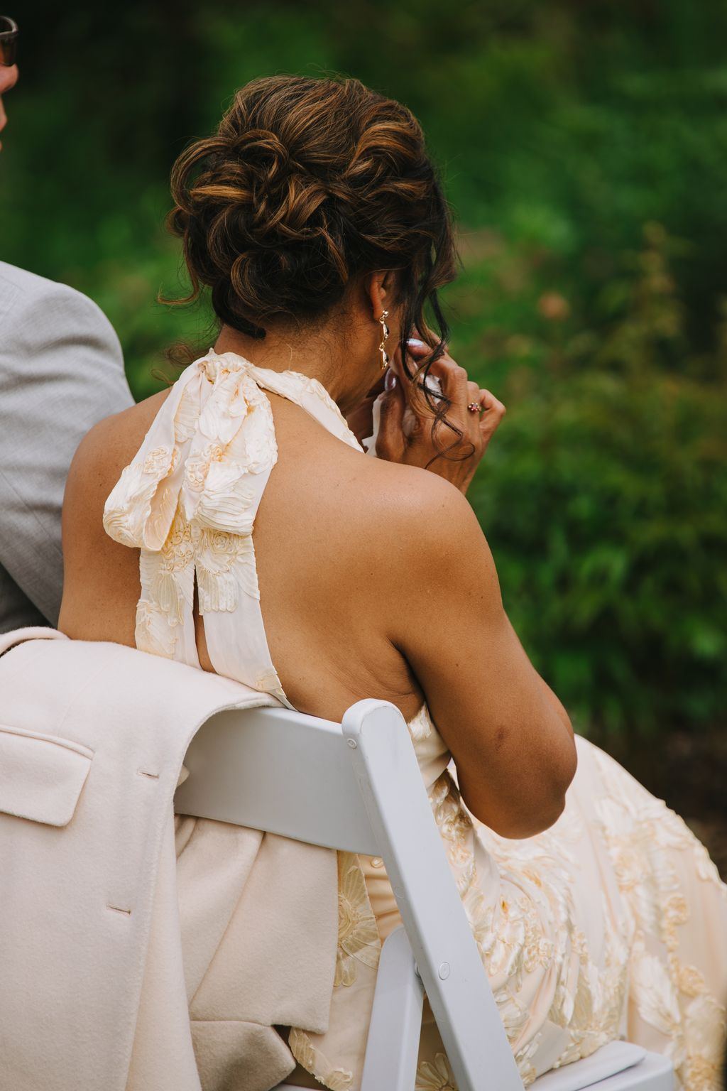 Bride from behind in elegant cream wedding dress with ornate beading, hair styled in romantic updo with loose curls, seated outdoors against soft green backg…