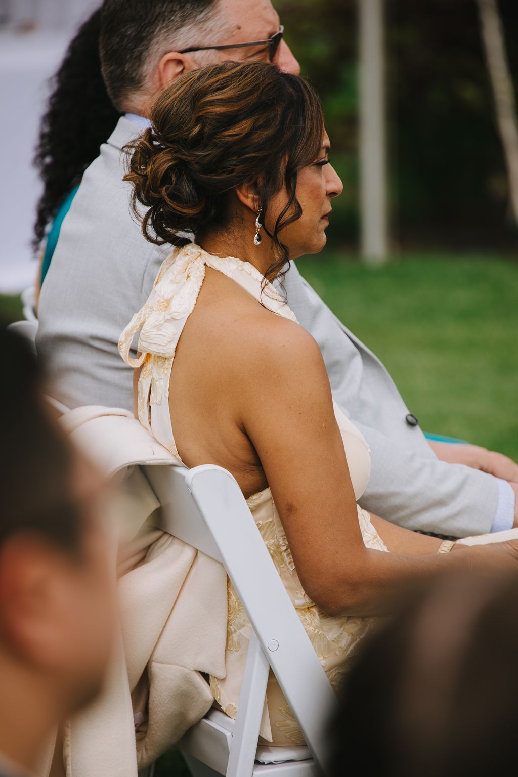 Bride in cream halter gown with upswept hair stands beside groom in profile during outdoor wedding ceremony