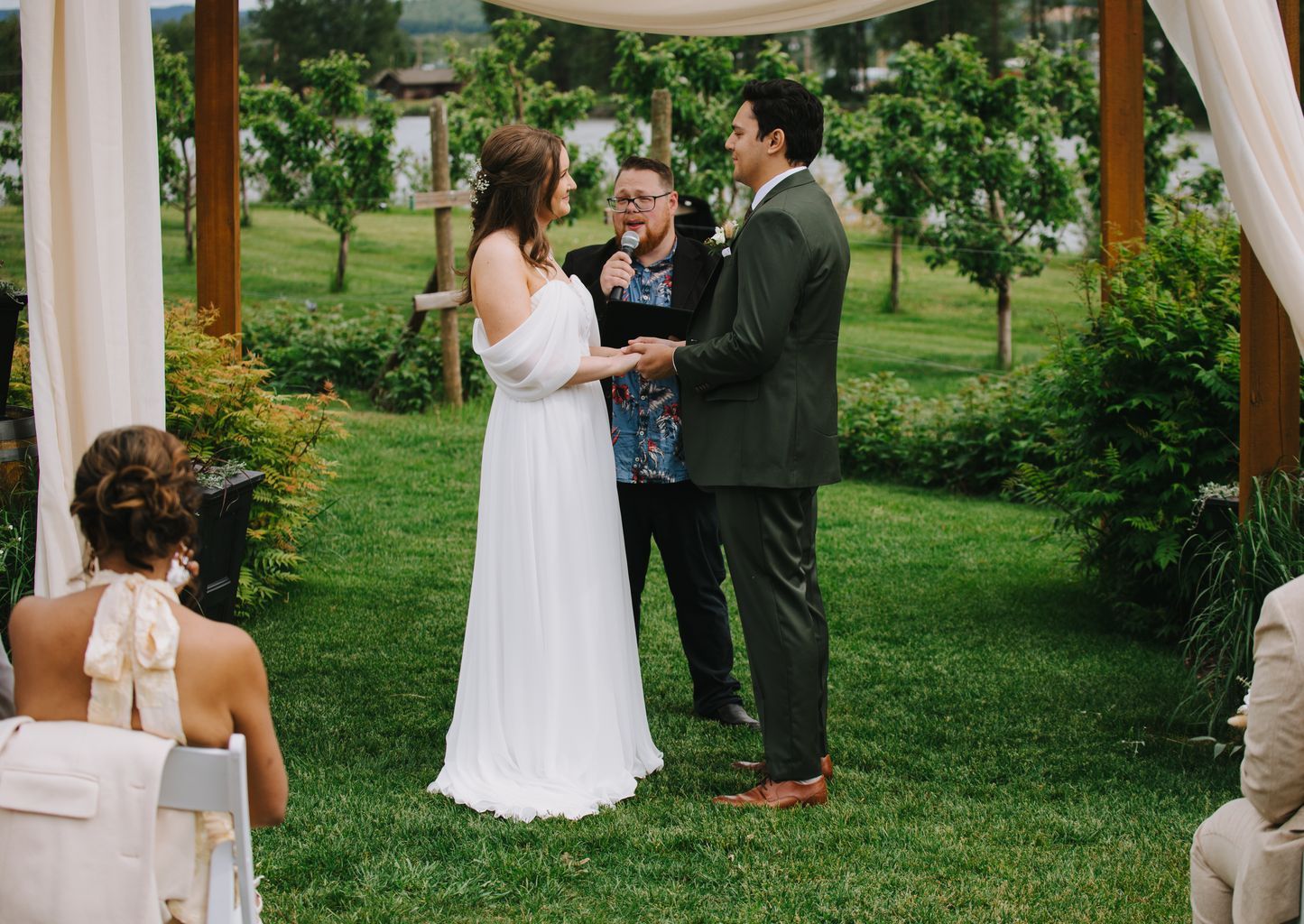 Bride in white off-shoulder gown and groom in olive suit exchange vows with officiant under ivy-covered pergola in lush garden setting