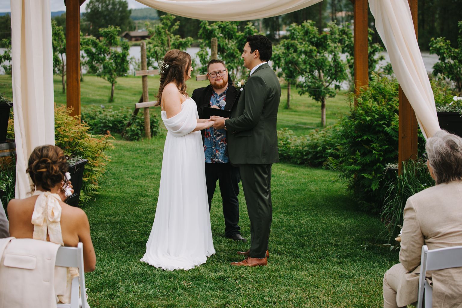 Bride in flowing white dress and groom in olive suit exchange vows with officiant under arbor in sunlit garden ceremony
