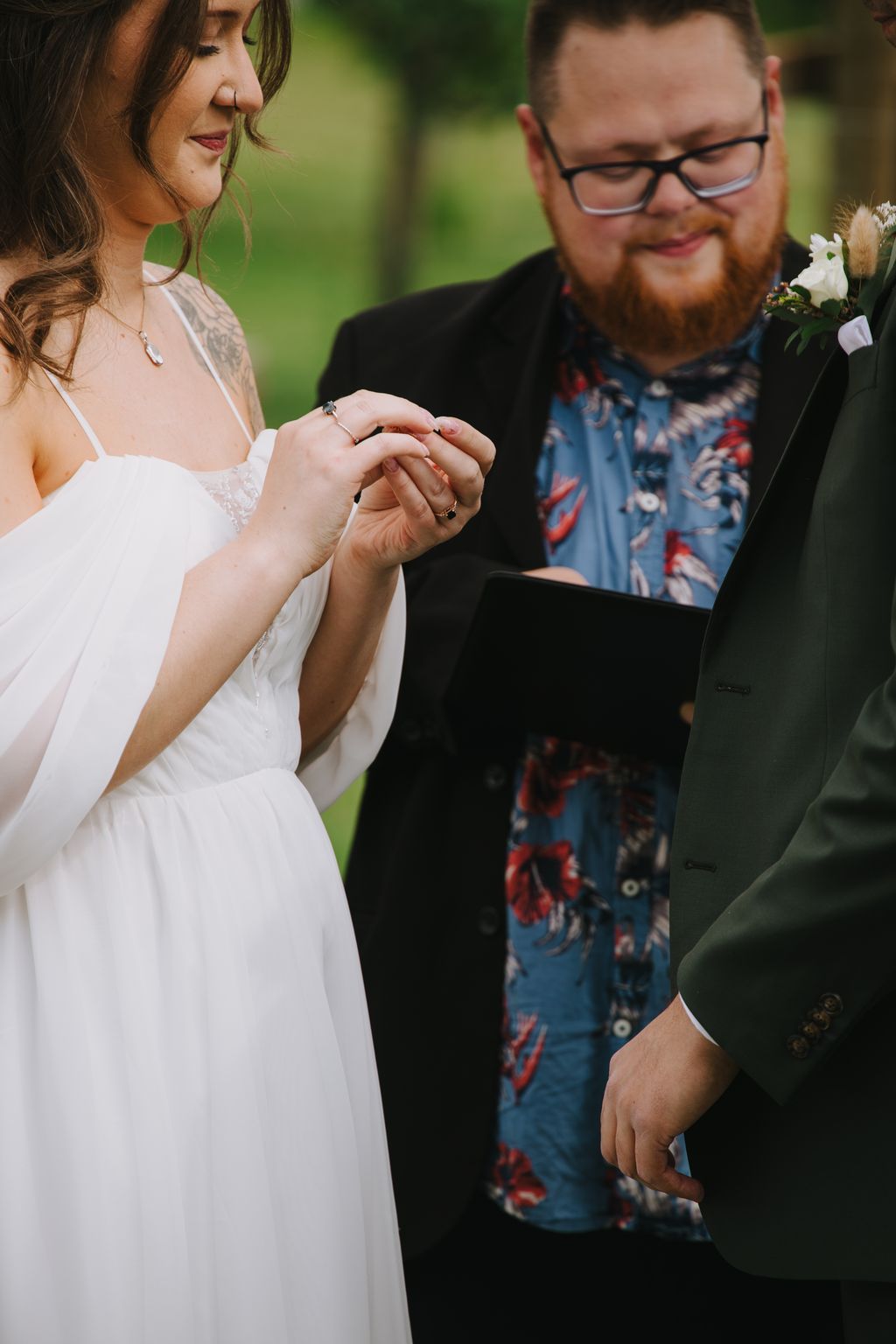 Bride and groom exchange rings outdoors with soft green background, bride in white dress and groom in floral shirt wearing glasses