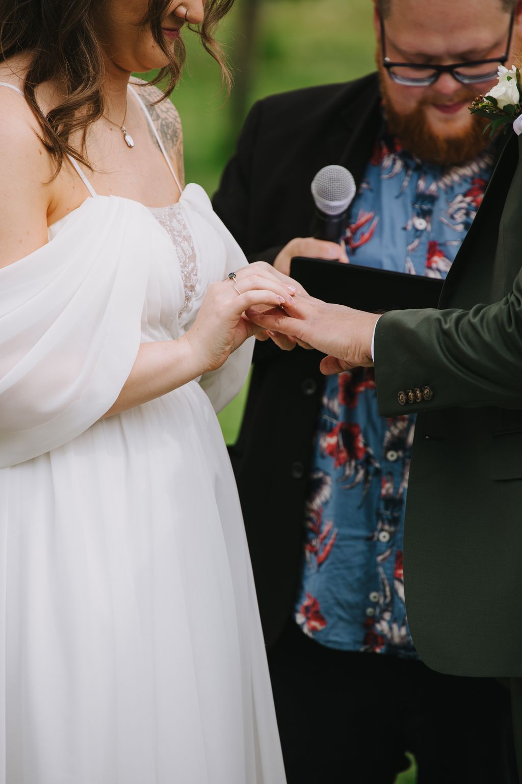 Bride in white lace dress placing wedding ring on groom''s finger during outdoor ceremony with green background