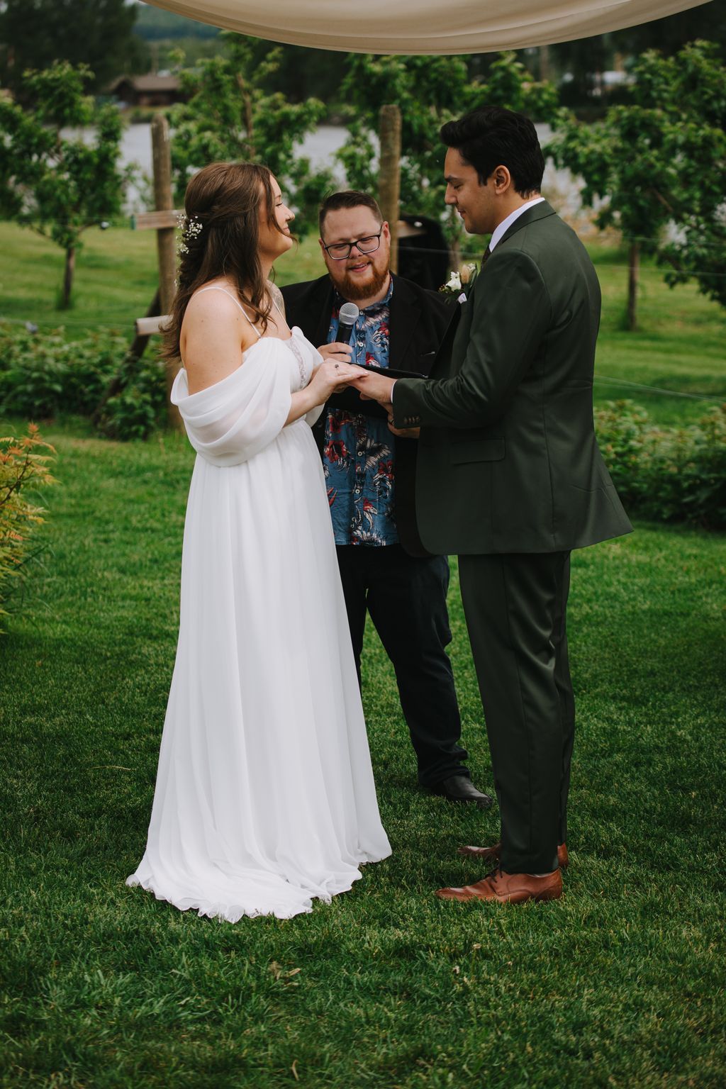 Bride and groom exchange rings during outdoor ceremony with officiant holding microphone under gazebo, lush green backdrop