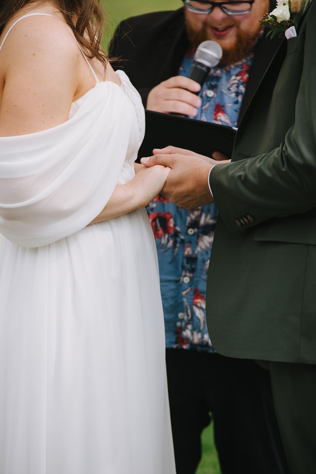Bride in white dress holding groom''s hand during outdoor ceremony with officiant holding microphone in background