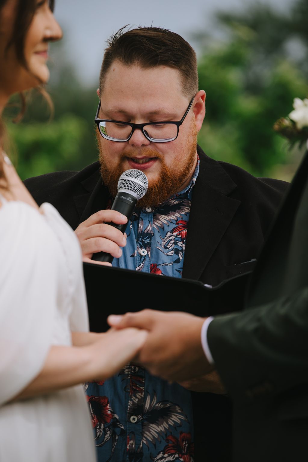 Bearded man with glasses speaks into microphone during outdoor ceremony, holding tablet, with blurred green background