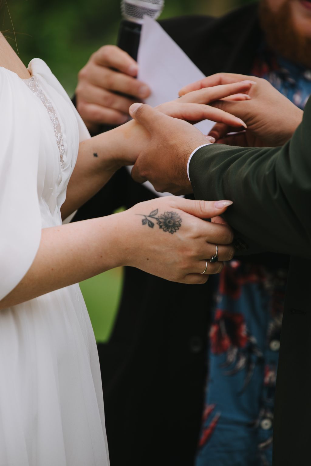 Bride and groom''s hands joined together displaying wedding rings and her sunflower tattoo in natural outdoor light