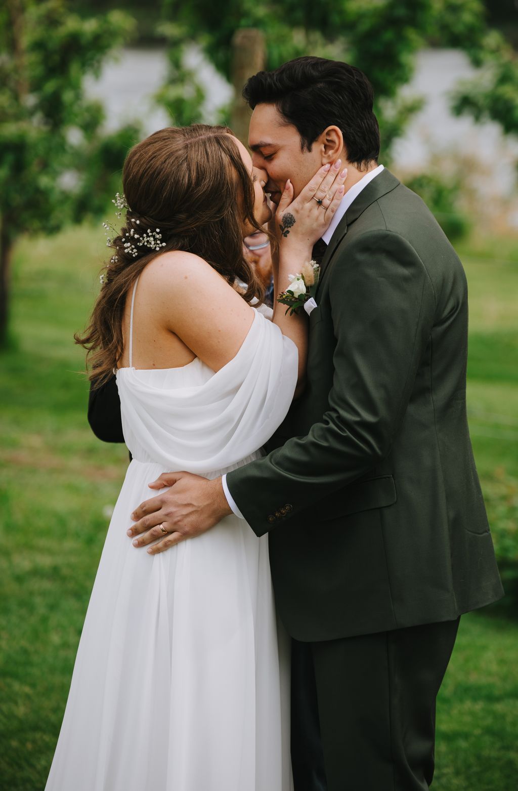 Bride and groom kiss outdoors, bride in white draped gown with pearl hair pins, groom in sage green suit, lush green garden background