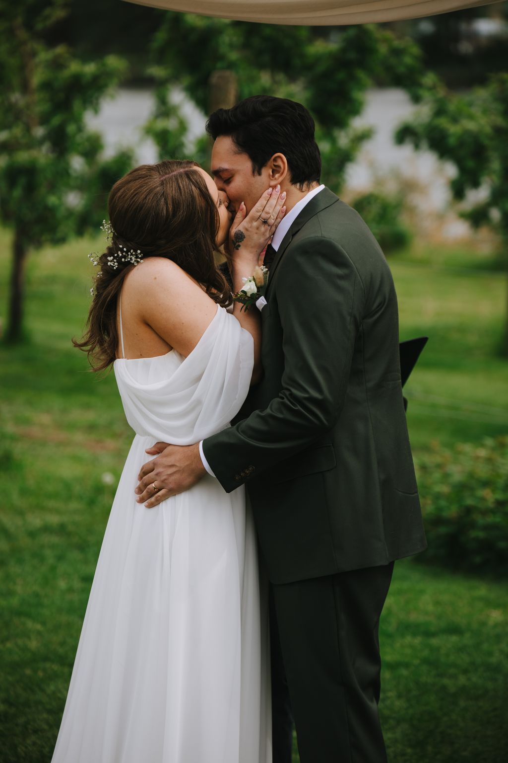 Bride and groom share a tender kiss beneath a wooden pergola with lush green foliage and white flowers in soft natural light