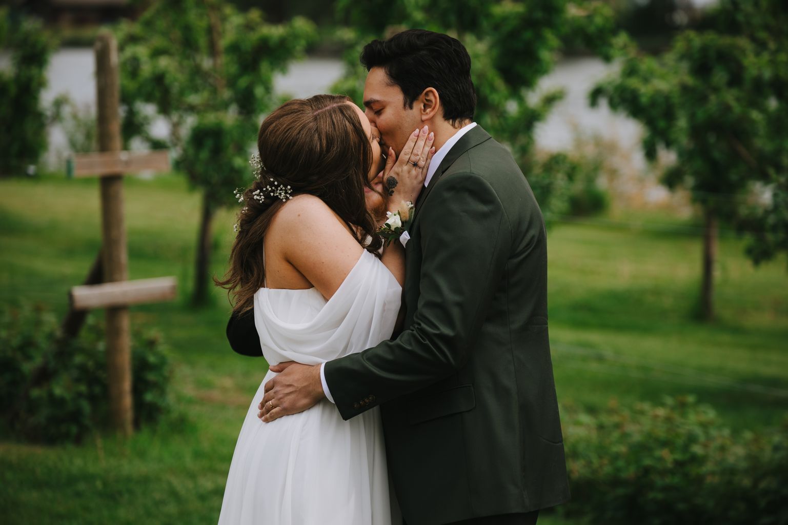 Bride in white draped gown and groom in olive suit kiss outdoors with lush green foliage behind them