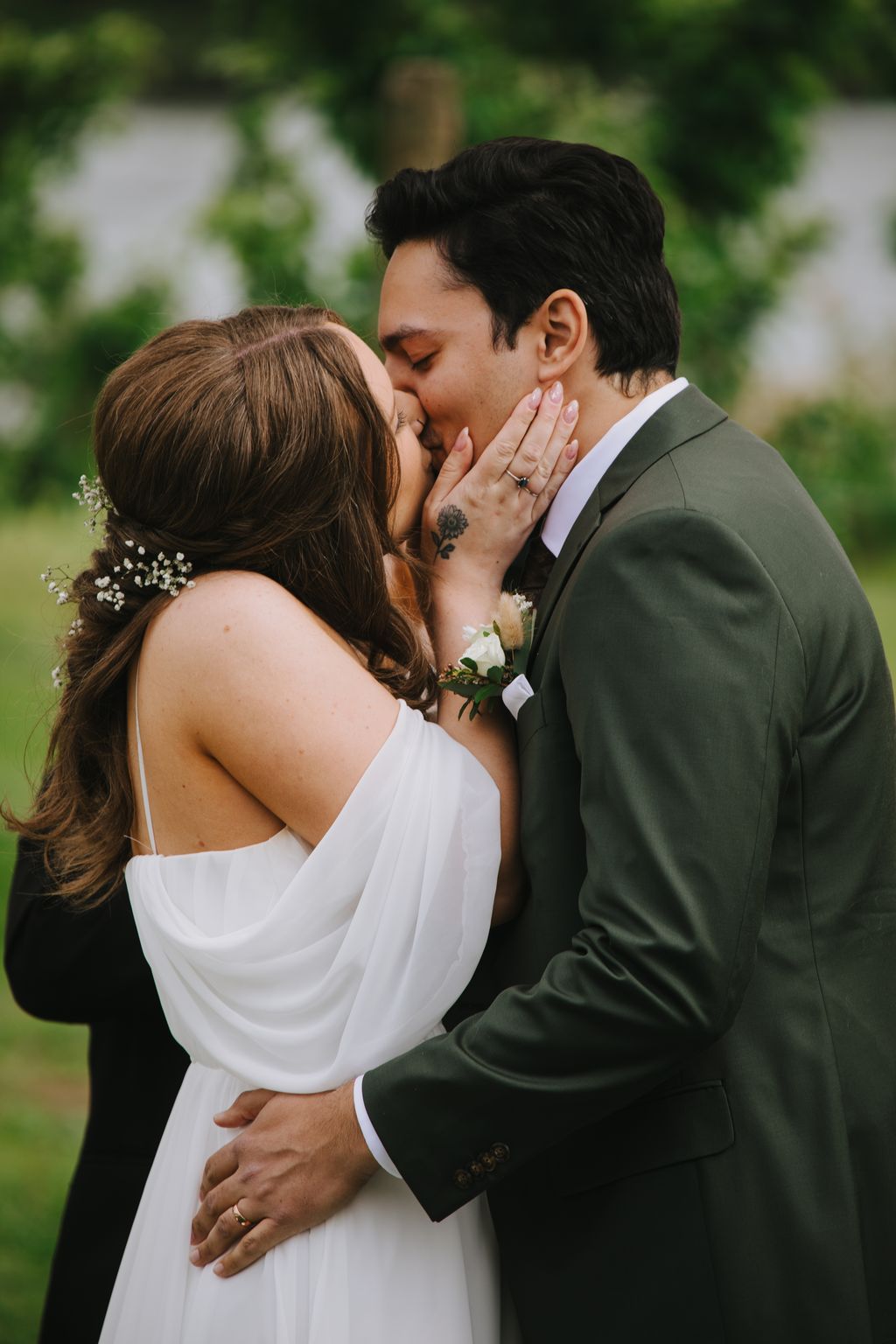 Bride and groom kiss outdoors, bride in white dress displaying ring, groom in sage suit, soft natural light with blurred green background