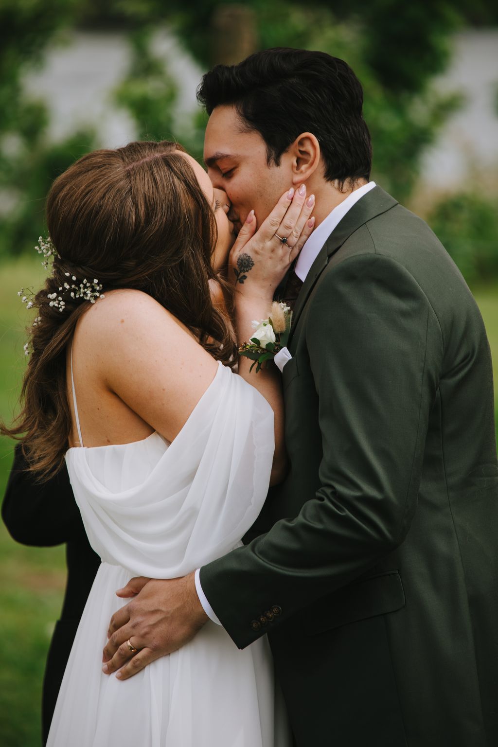 Bride and groom kissing outdoors, bride in white dress holding bouquet, groom in sage green suit, soft natural light with blurred green background