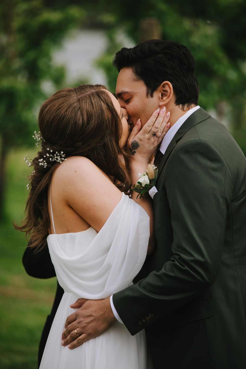 Bride in white off-shoulder dress and groom in dark suit kiss tenderly outdoors with blurred green foliage background