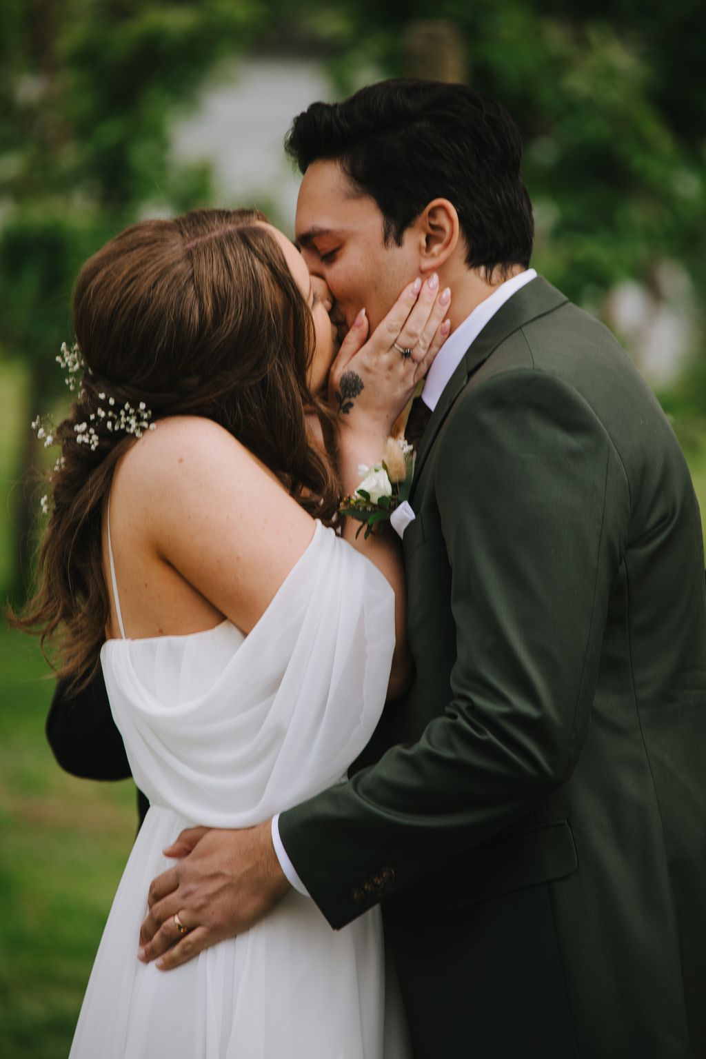 Bride and groom kiss outdoors in soft natural light, bride in white dress with bouquet, groom in olive suit with ring visible