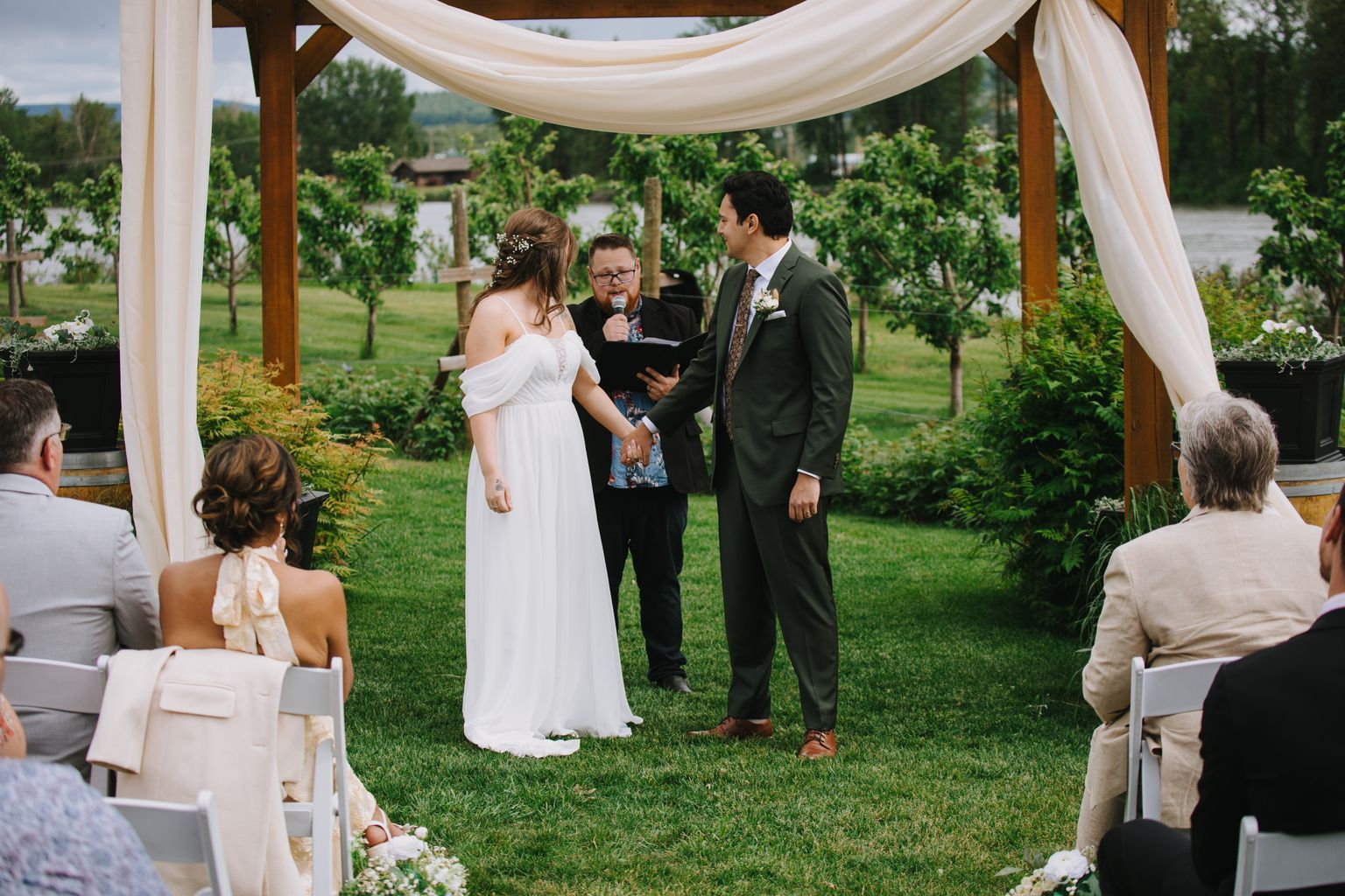 Bride in white off-shoulder gown and groom in olive suit exchange vows under draped fabric canopy with officiant, manicured garden backdrop