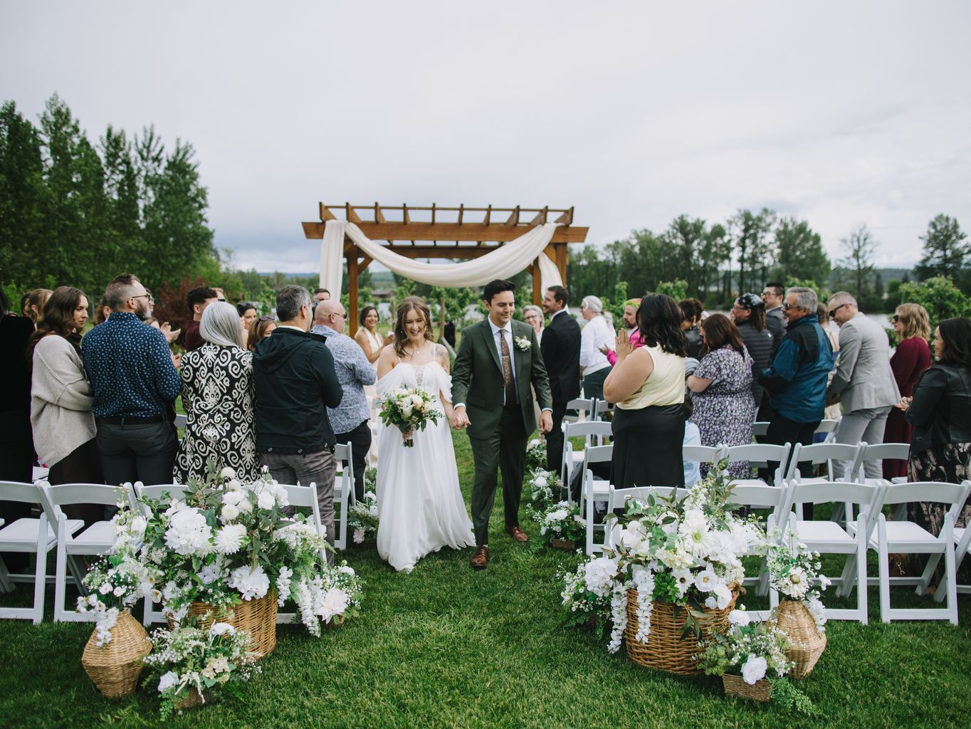 Bride and groom walk down outdoor aisle under wooden pergola with white fabric draping and floral arrangements on a sunny day