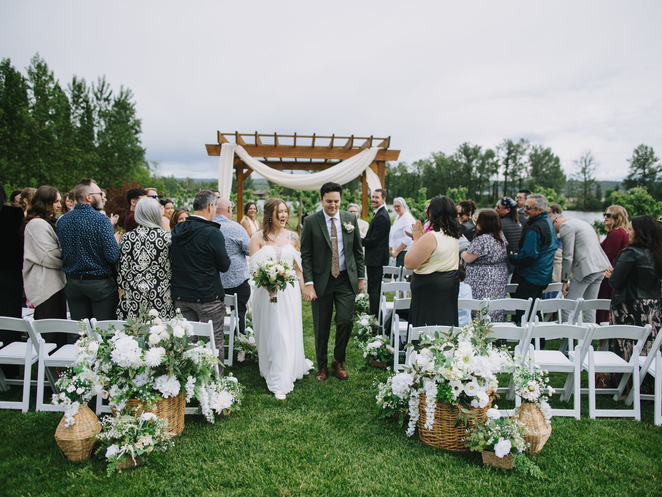 Bride and groom walk down outdoor aisle under wooden pergola with white draping and floral arrangements on grass