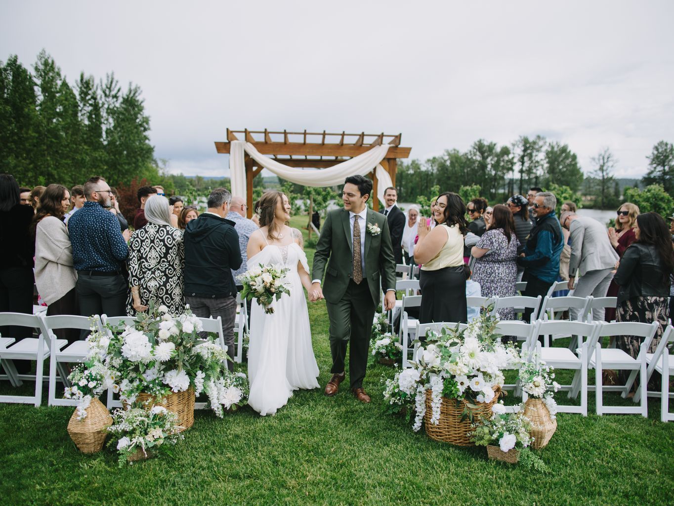 Bride and groom walk hand in hand down grassy aisle under wooden pergola with flowing white fabric at outdoor garden wedding ceremony