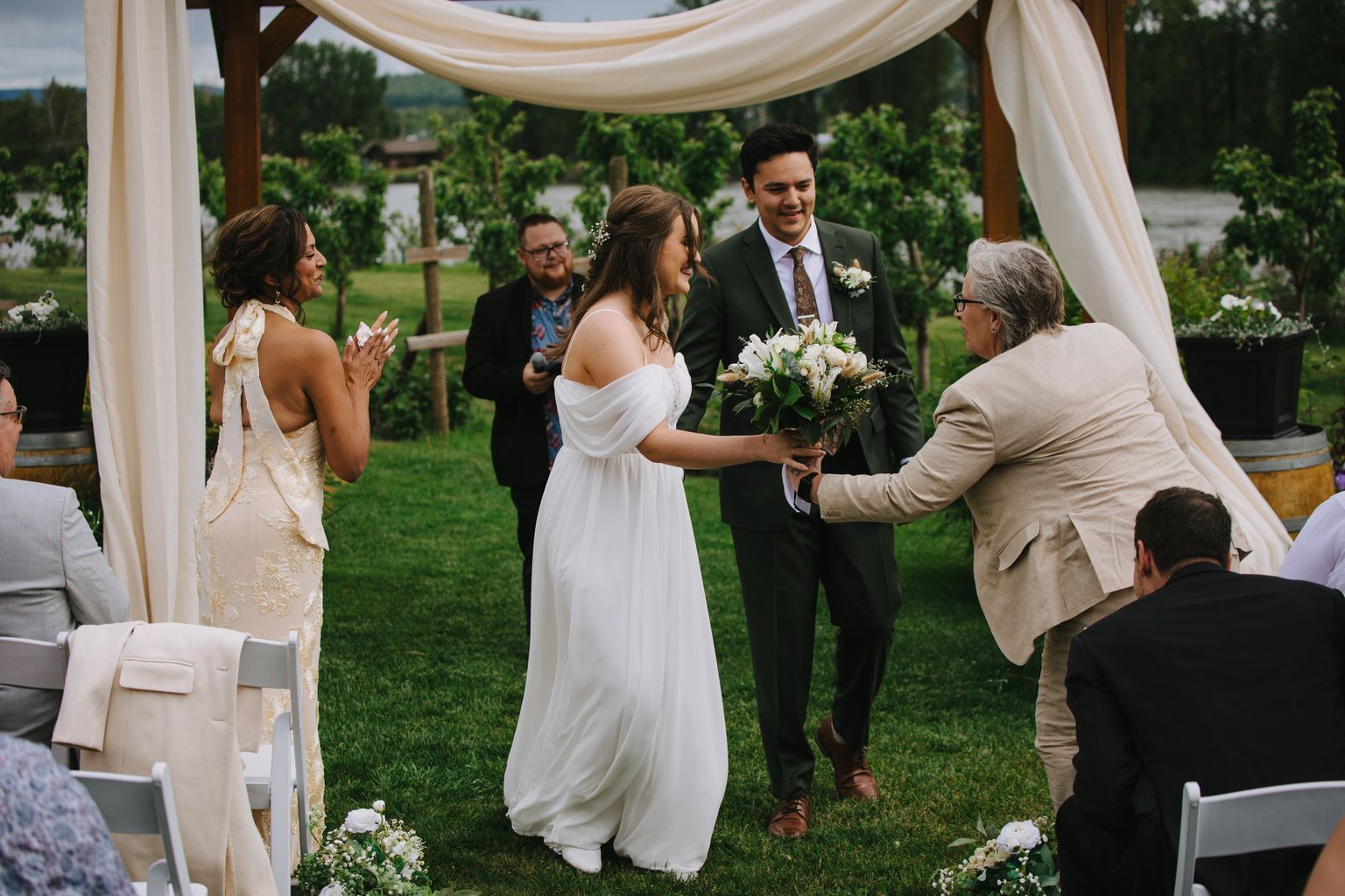 Bride in flowing white off-shoulder gown holds bouquet while exchanging vows with groom in dark suit during outdoor garden ceremony under cream fabric canopy