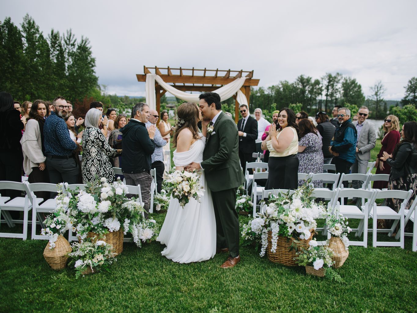 Bride and groom kiss under wooden pergola with white draping and floral arrangements at outdoor garden ceremony